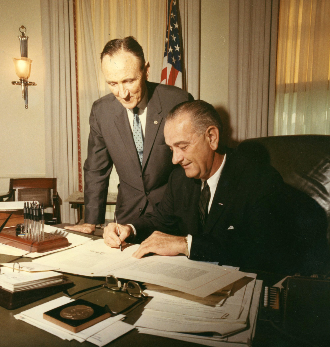 Senator Mike Mansfield leaning over President Lyndon Johnson who is seated at a typewriter.