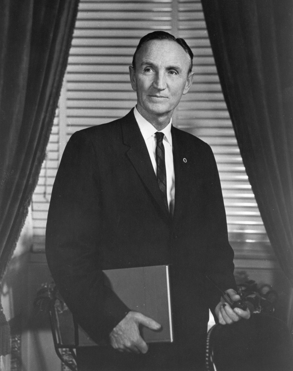 Senator Mike Mansfield standing and holding a book and his pipe.