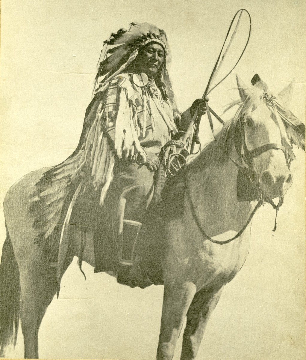 Alaxchíiahush wearing his war shirt decorated with ermines and a feathered headdress, sitting astride a white horse.