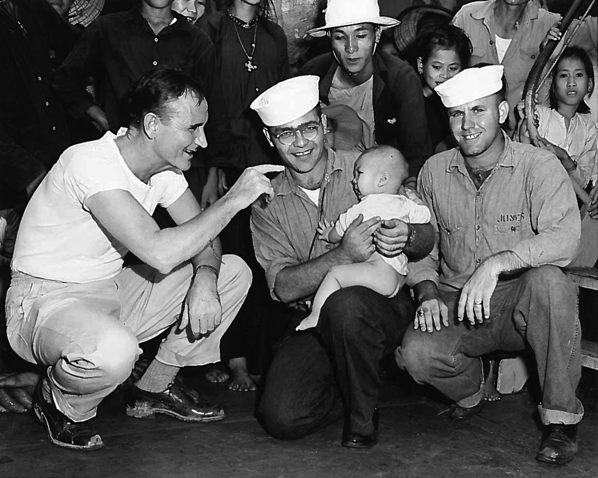 Senator Mansfield kneeling and smiling at an Vietnamese infant who is being held by a U.S. Navy serviceman.