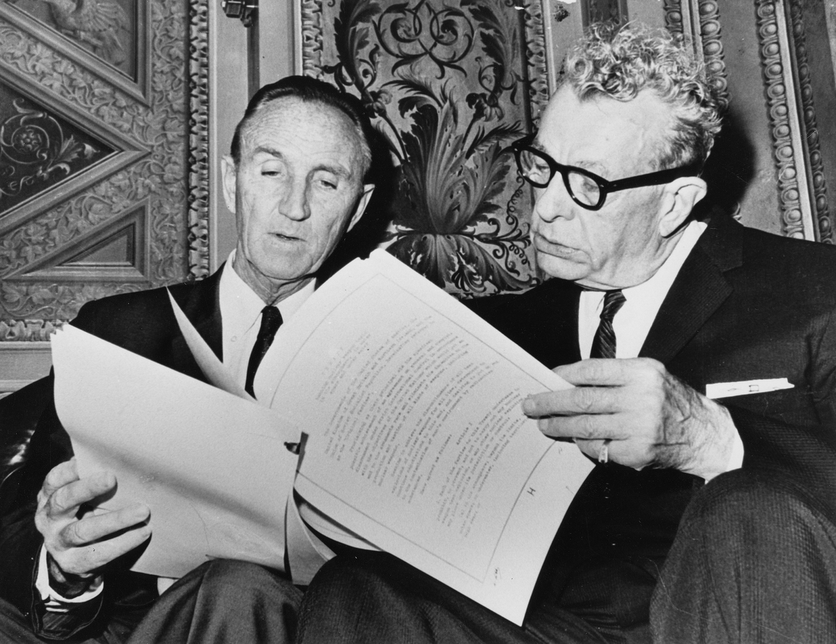 Senators Mike Mansfield and Everett Dirksen seated on a couch and reading from the same document.