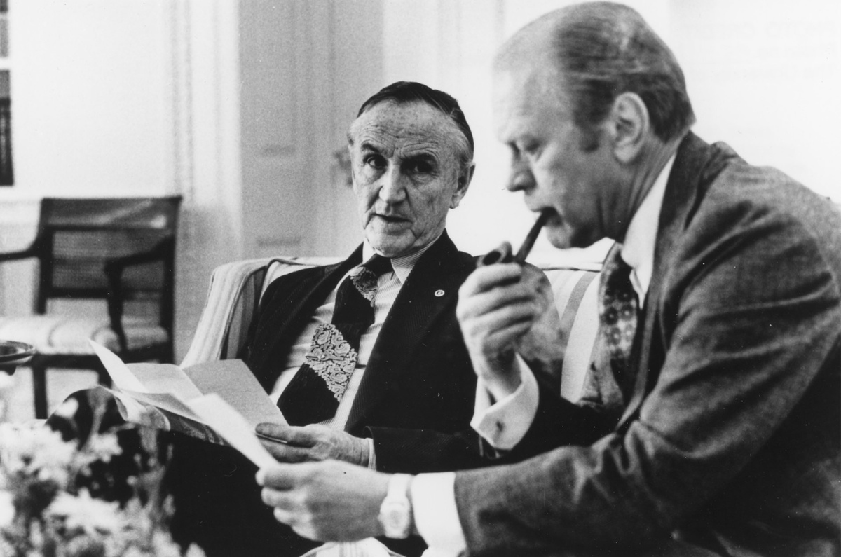 Senator Mike Mansfield seated on a couch with President Gerald Ford who is reading a document and smoking a pipe.