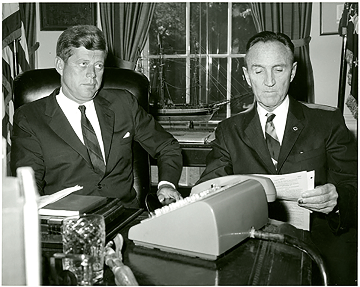 Senator Mike Mansfield and President John F. Kennedy sitting next to each other at a desk with a typewriter in front of them.
