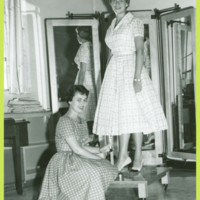 Two women pose while measuring a hemline.